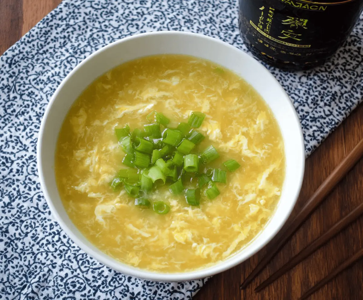 A bowl of hot Egg Drop Soup with wispy egg ribbons and chopped green onions on top, served in a white bowl with a spoon.
